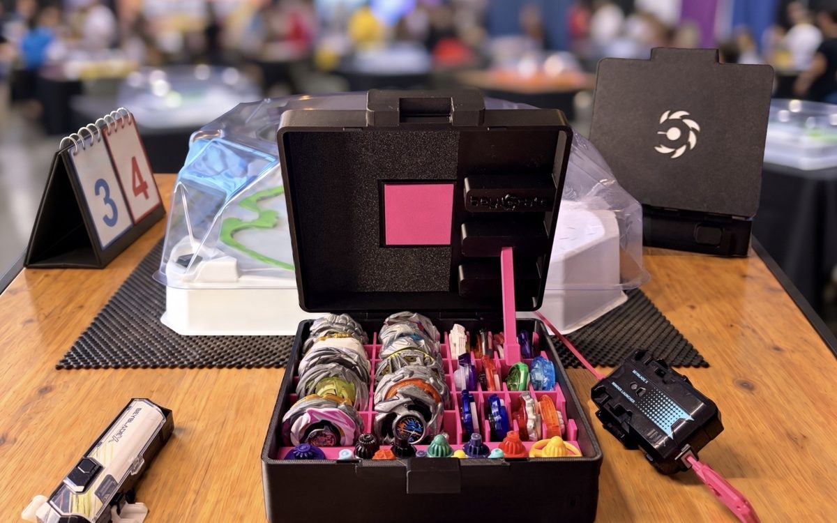 The Beycrate 1 open on a tournament table with the Tokyo Pink insert, loaded with blades, ratchets and bits, next to a Beyblade X stadium, launchers and a scoreboard